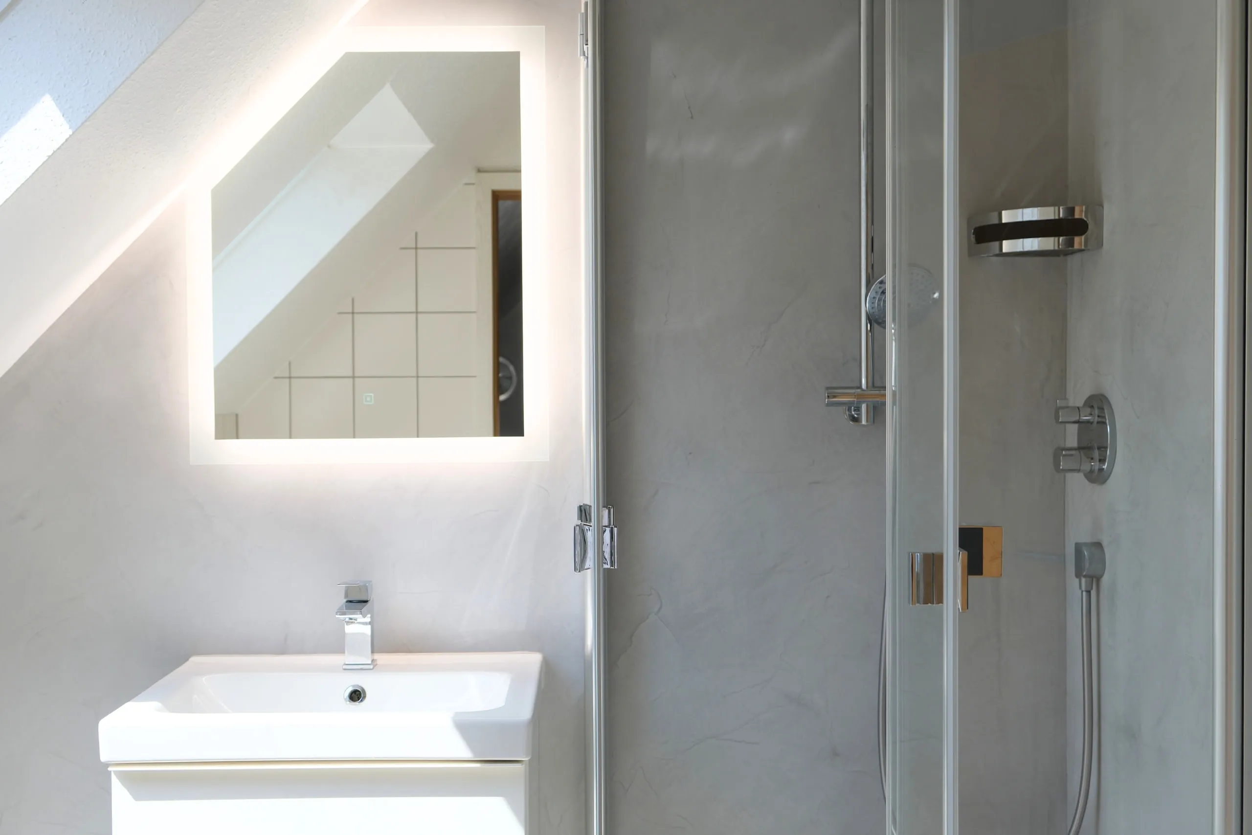 Waschtisch unter beleuchtetem Spiegel in der Dachschräge, daneben eine Dusche mit Glasabtrennung